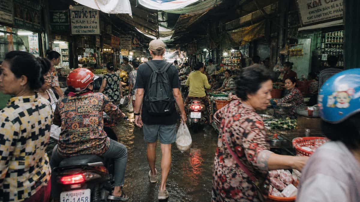 Allée de marché couvert en Asie du Sud-Est avec étals frais, sol humide et circulation dense entre habitants et voyageurs